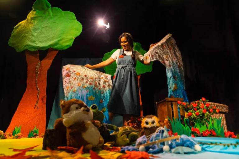 Paloma Mariscal en escena moviendo telas pintadas que representan el bosque, rodeada de títeres de animales